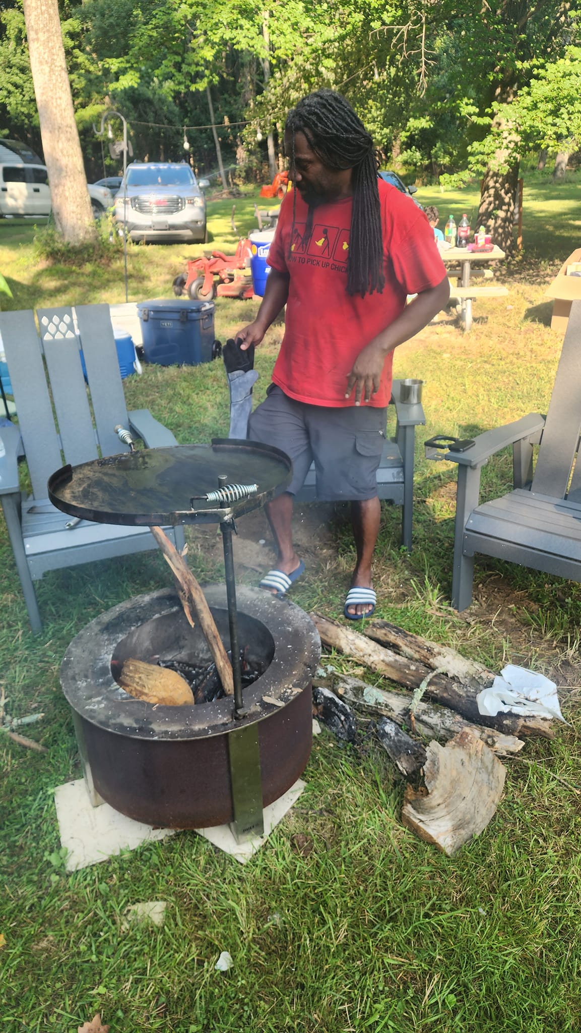 Tending the fire pit grill in a red shirt with long locs at the first campout — Campout 2023