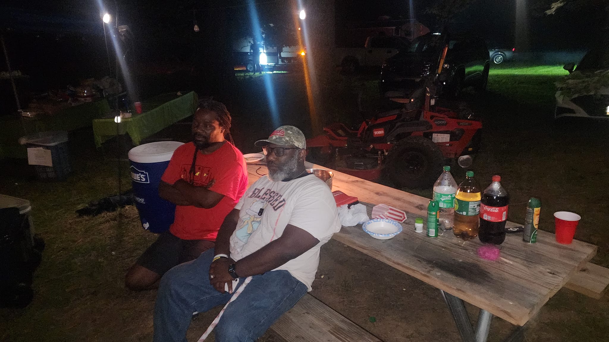 Two guests relaxing at a picnic table at night with drinks and snacks — Campout 2023