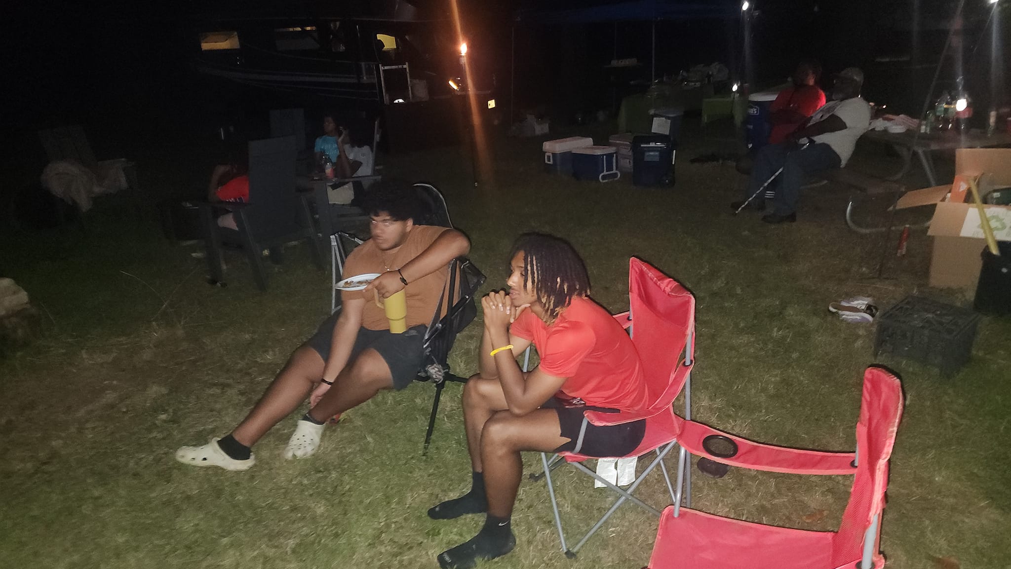 Two teens sitting in camp chairs at night eating and hanging out around the campsite — Campout 2023