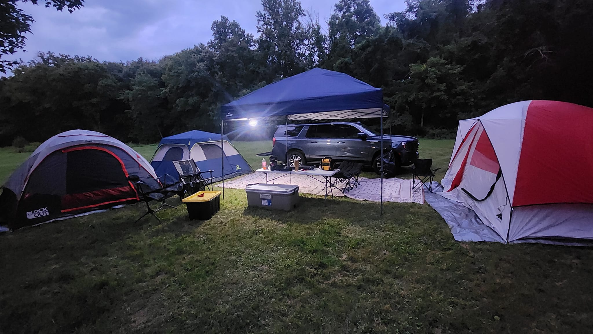Multiple tents and the SUV with canopy lit up on the open lawn at dusk — Campout 2023