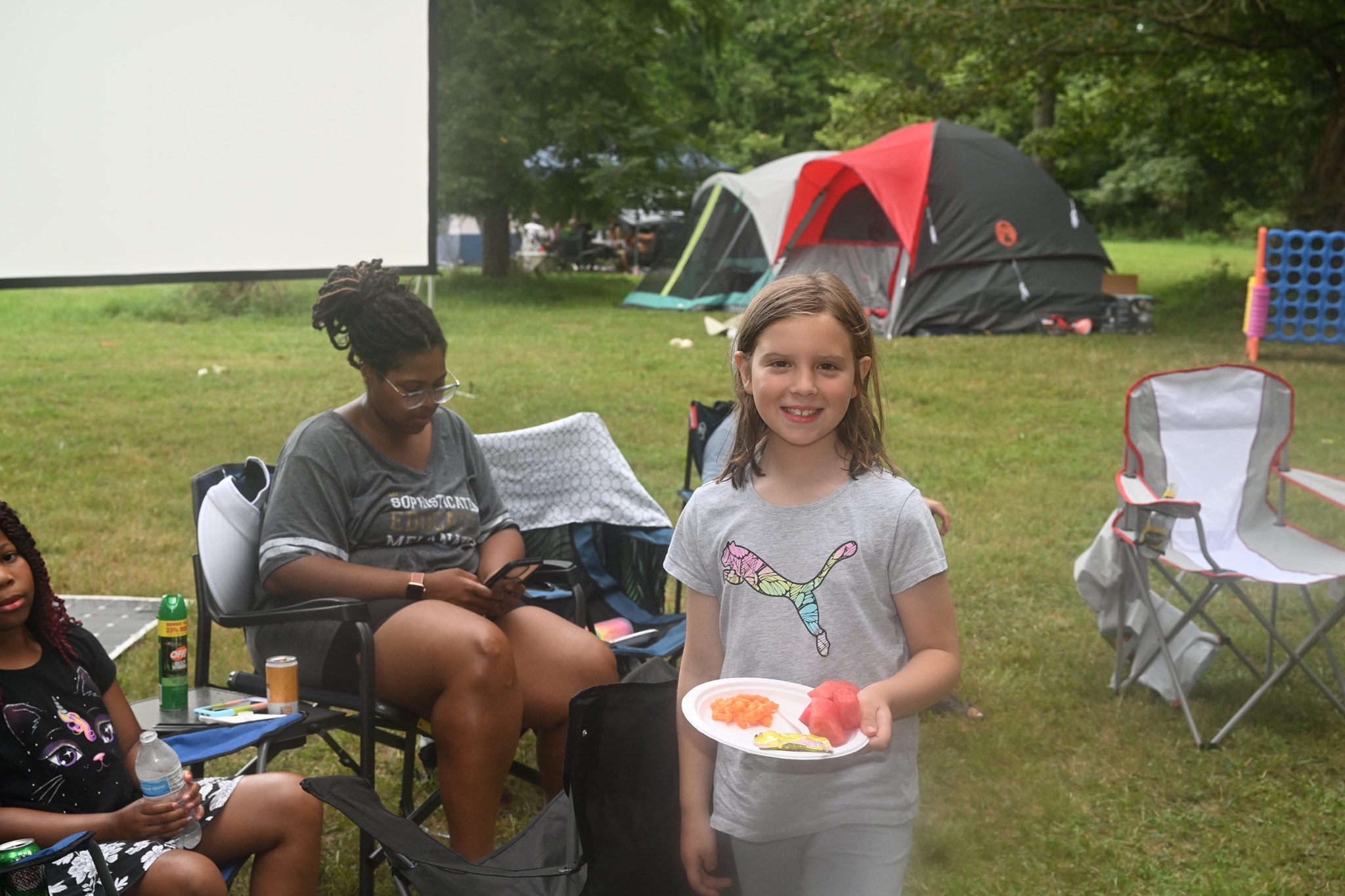 A girl holding a plate of food and smiling with the movie screen and tents behind her — Campout 2023