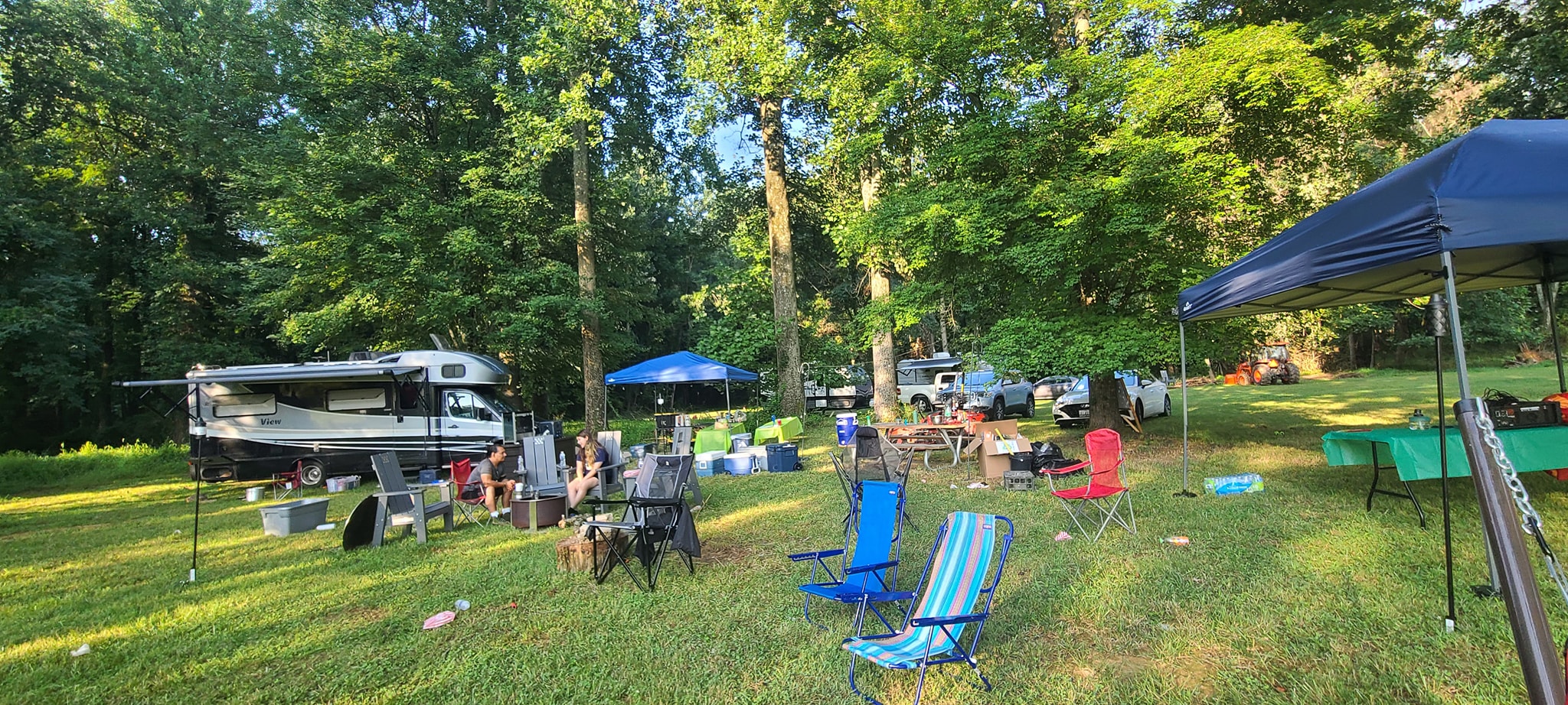 Wide view of the campsite with the RV, canopy tents, and camp chairs spread across the shaded lawn — Campout 2023
