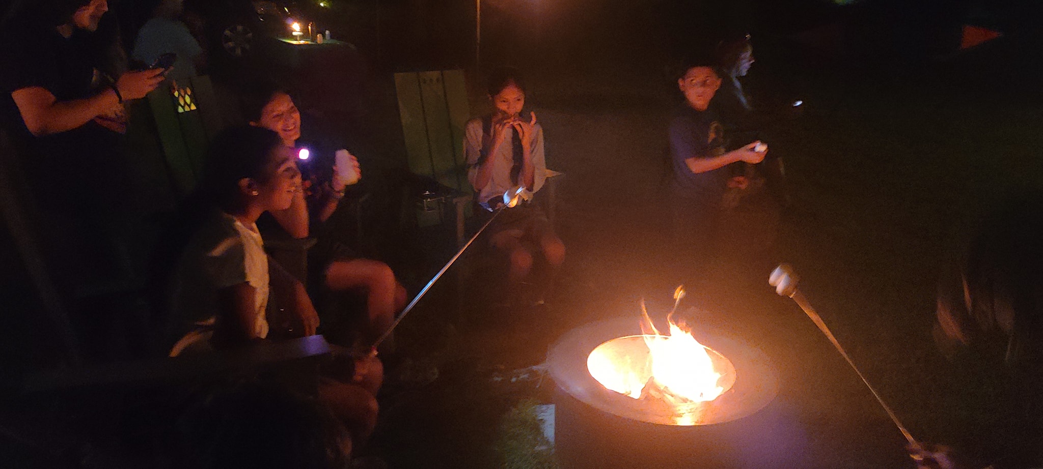 Kids roasting marshmallows over the fire pit at night — Campout 2023
