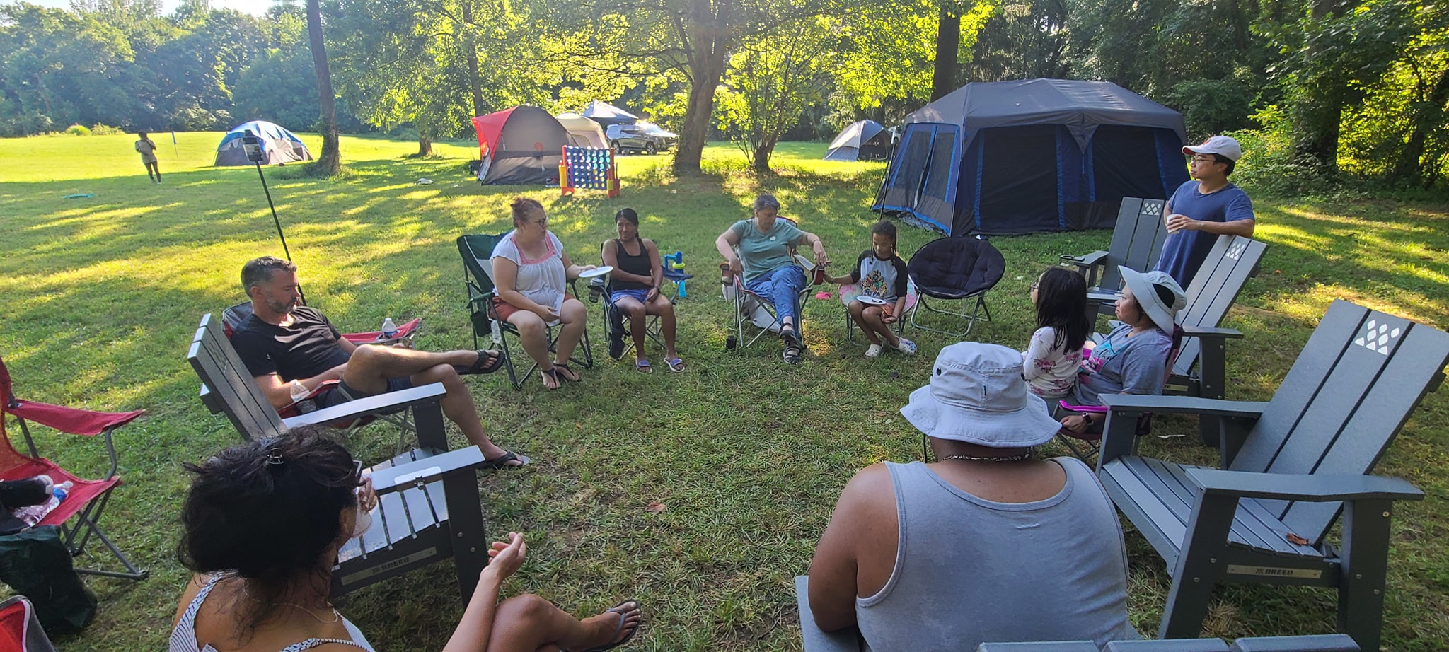 Guests sitting in a circle of camp chairs on the sunny lawn with tents behind — Campout 2023