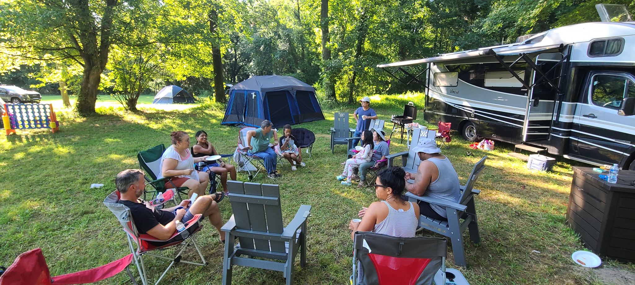 A wide circle of guests in camp chairs on the lawn beside the RV and tents — Campout 2023