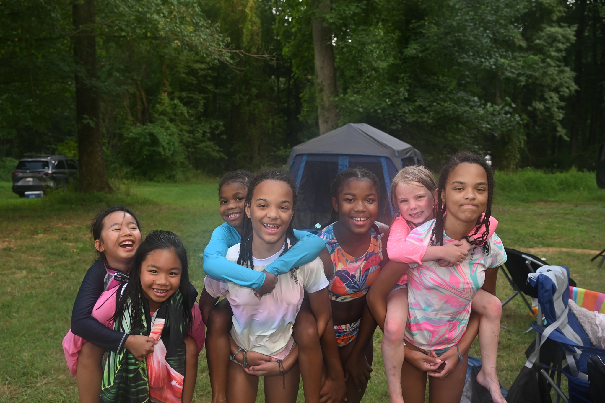 A group of girls smiling together in front of a tent in the wooded yard — Campout 2023