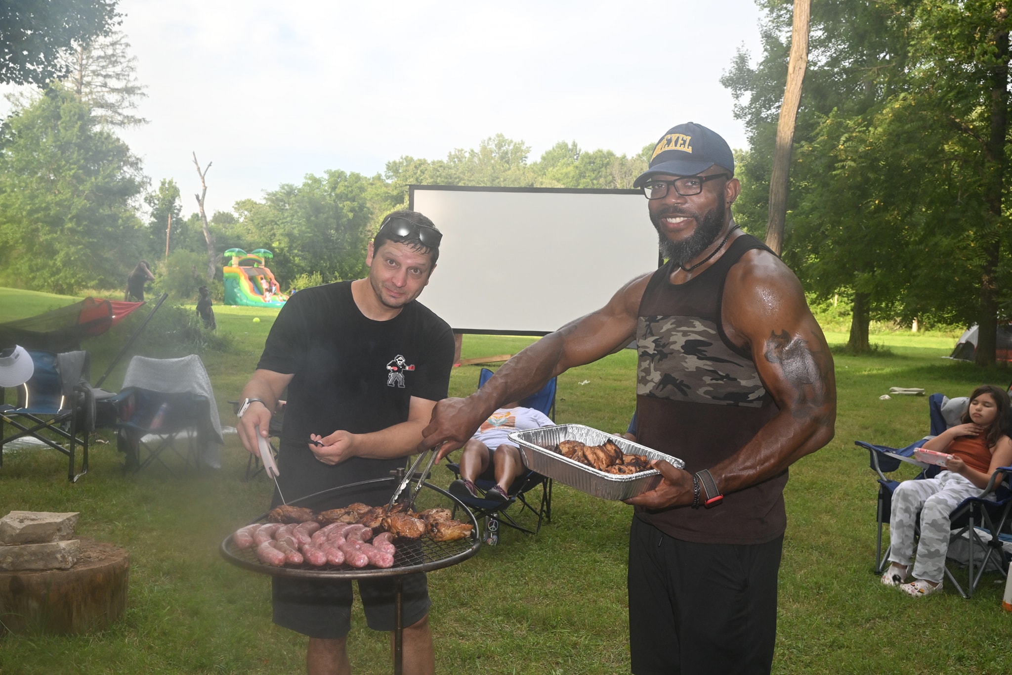 Two men grilling burgers and sausages with the movie screen and bounce house behind — Campout 2023