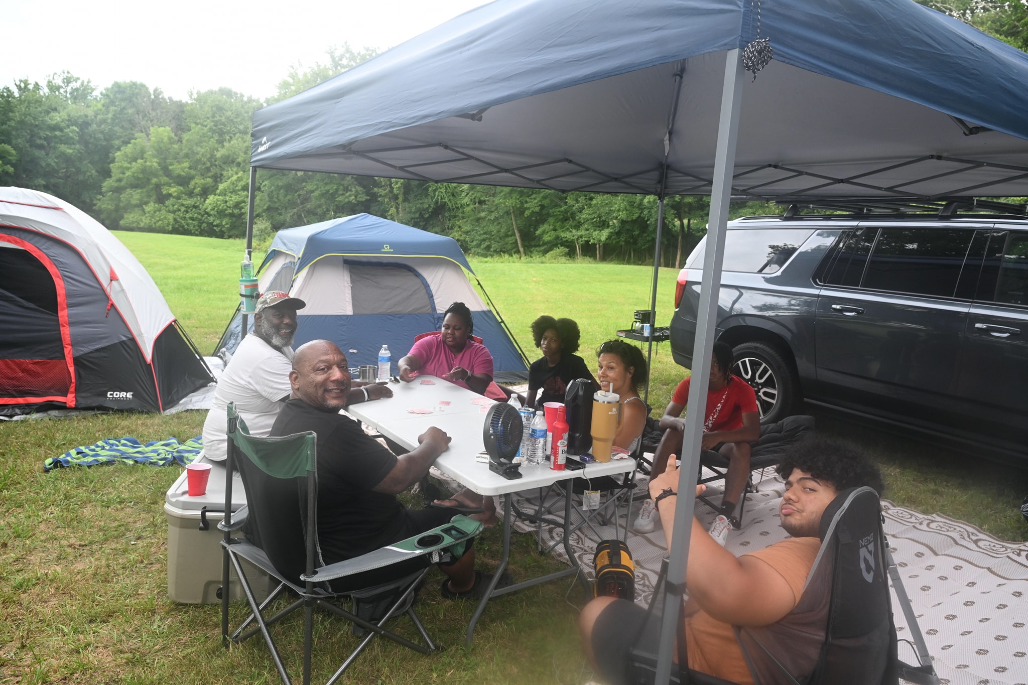A group playing cards together under the canopy with tents and the SUV nearby — Campout 2023