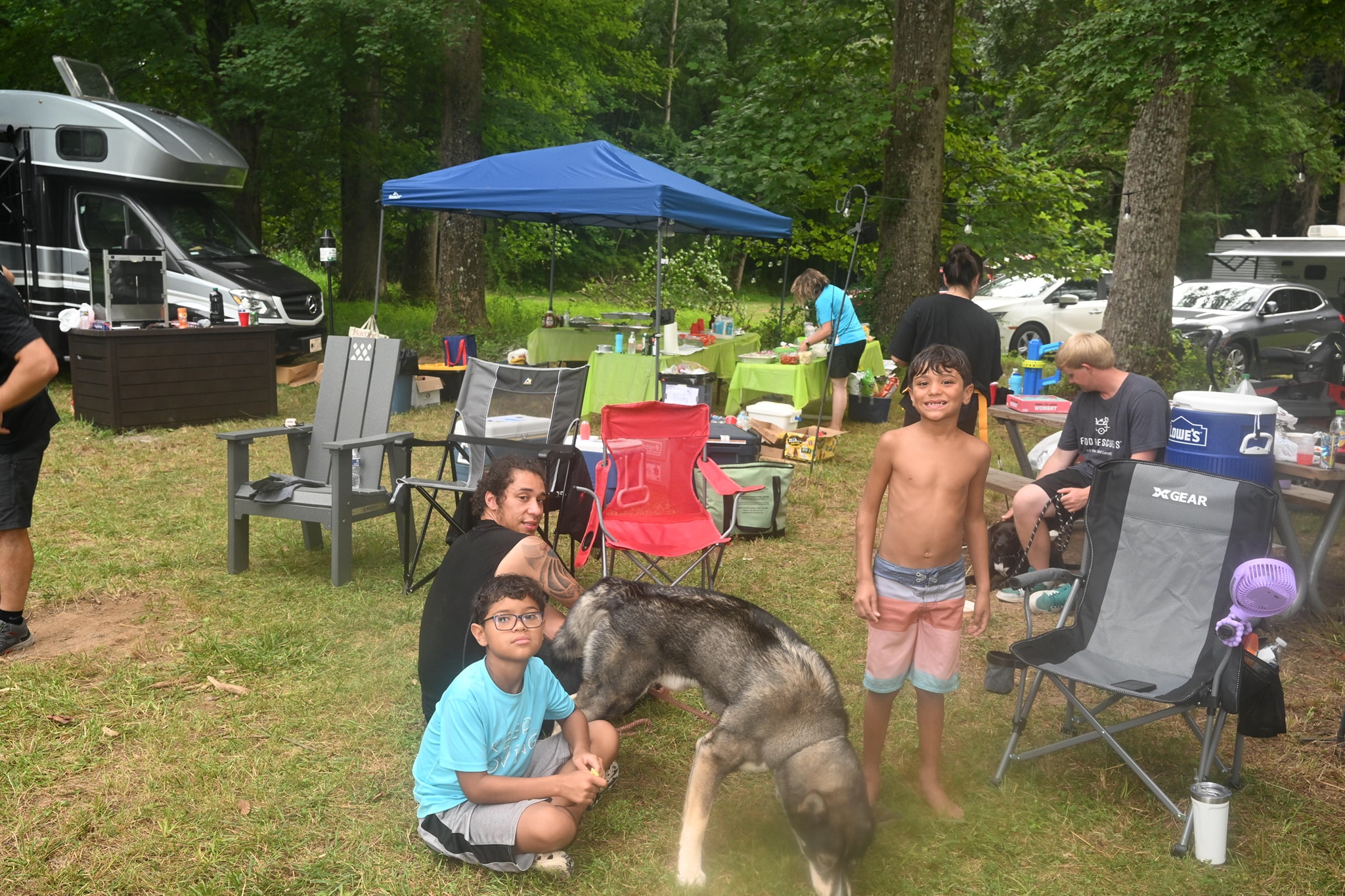 Kids playing with a large dog near the food station and RV in the wooded campsite — Campout 2023