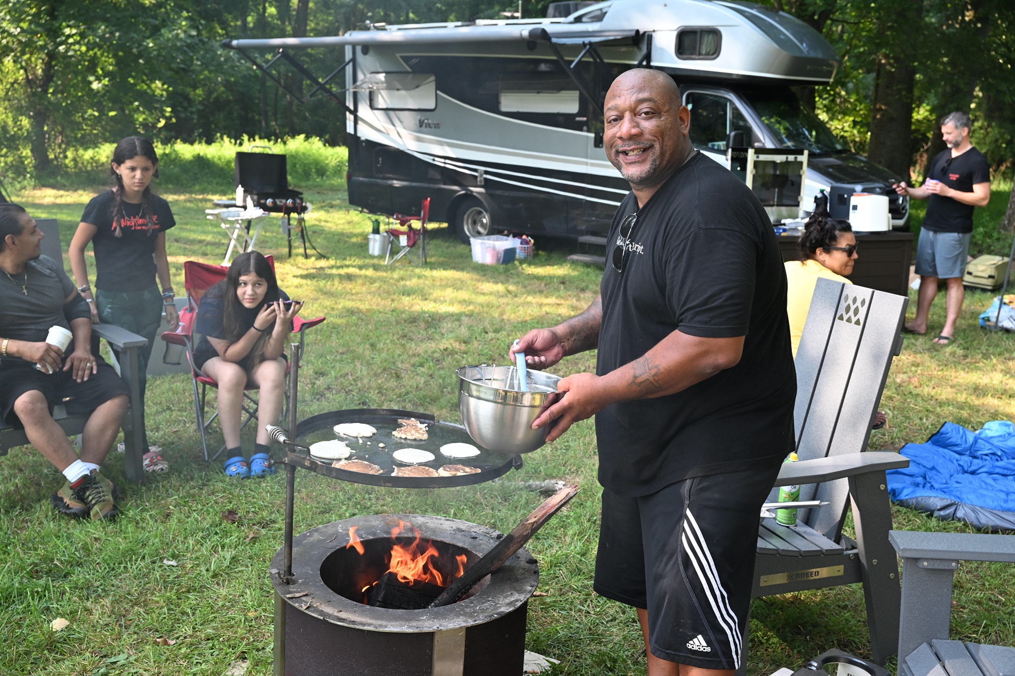Smiling and cooking burgers on the fire pit grill with guests relaxing behind — Campout 2023