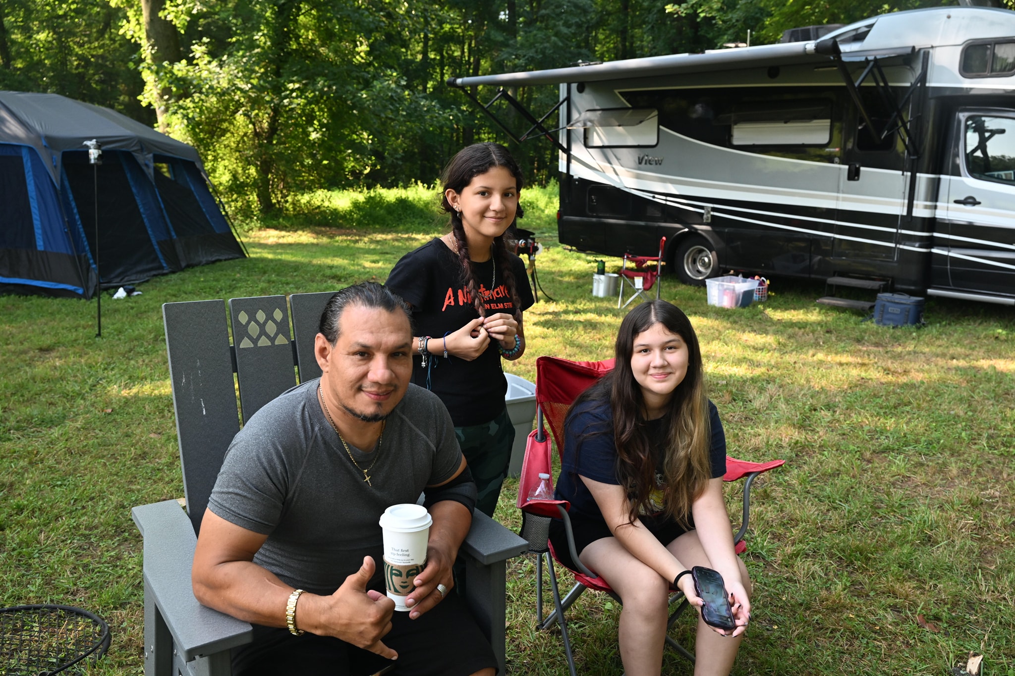 A dad and two daughters relaxing in Adirondack chairs beside the RV — Campout 2023