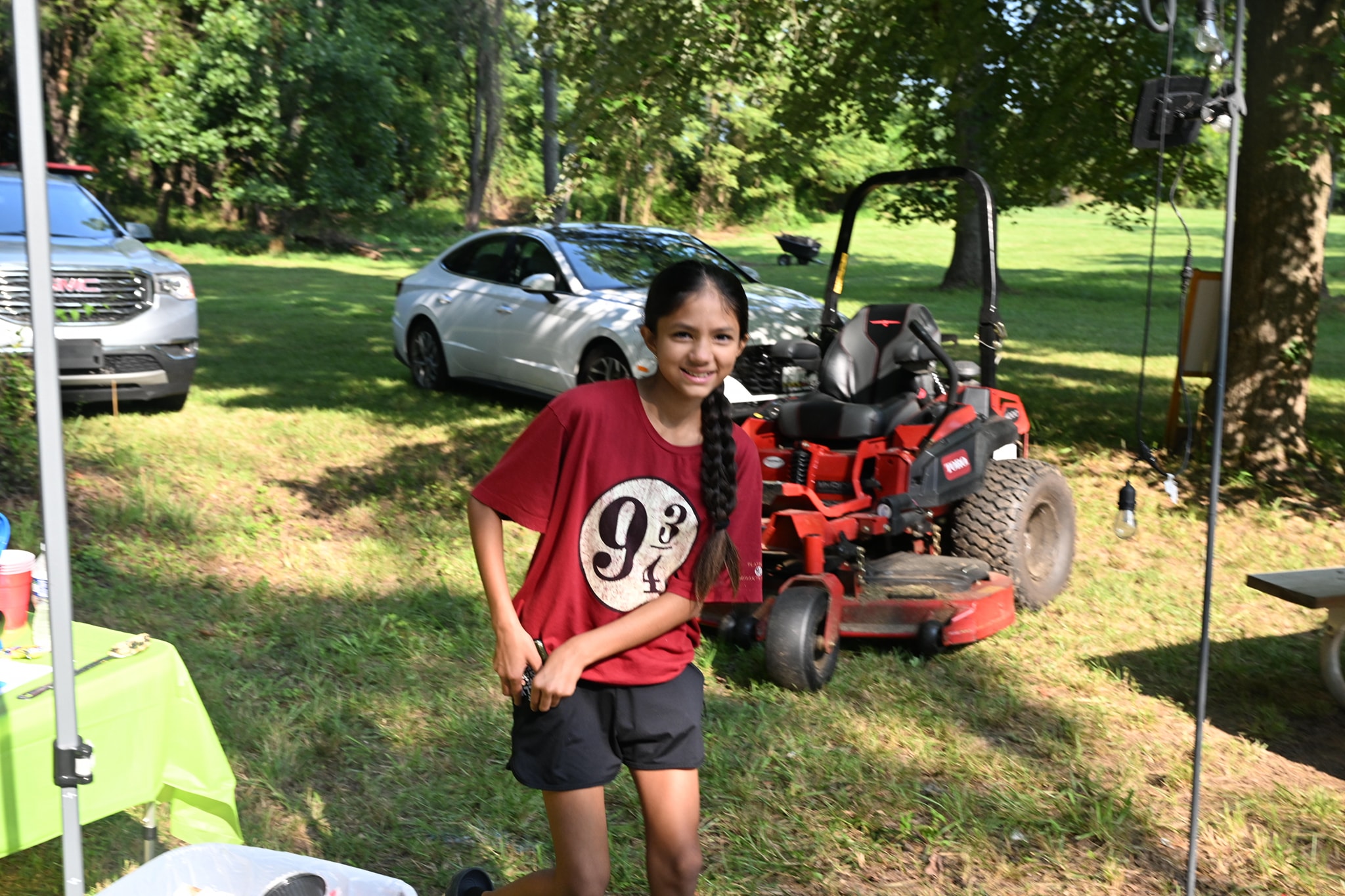 A girl in a Harry Potter Platform 9¾ shirt smiling beside the riding mower — Campout 2023