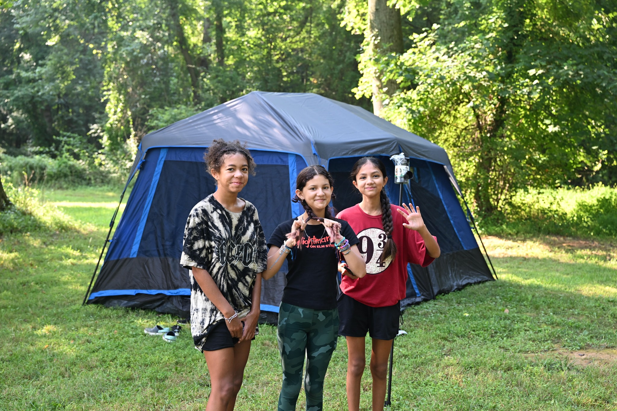 Three girls posing in front of a large tent in the wooded yard — Campout 2023