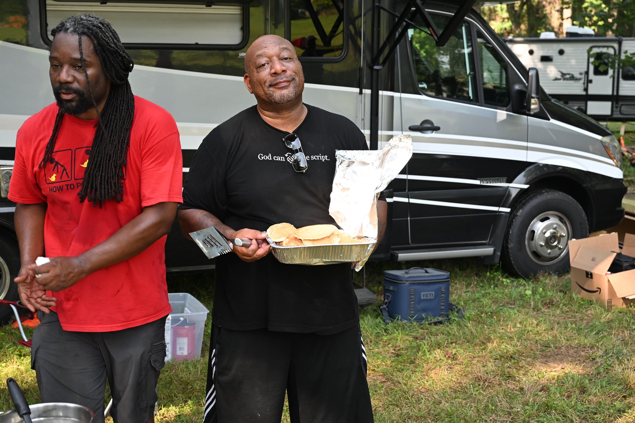Two men standing in front of the RV, one holding a tray of buns — Campout 2023