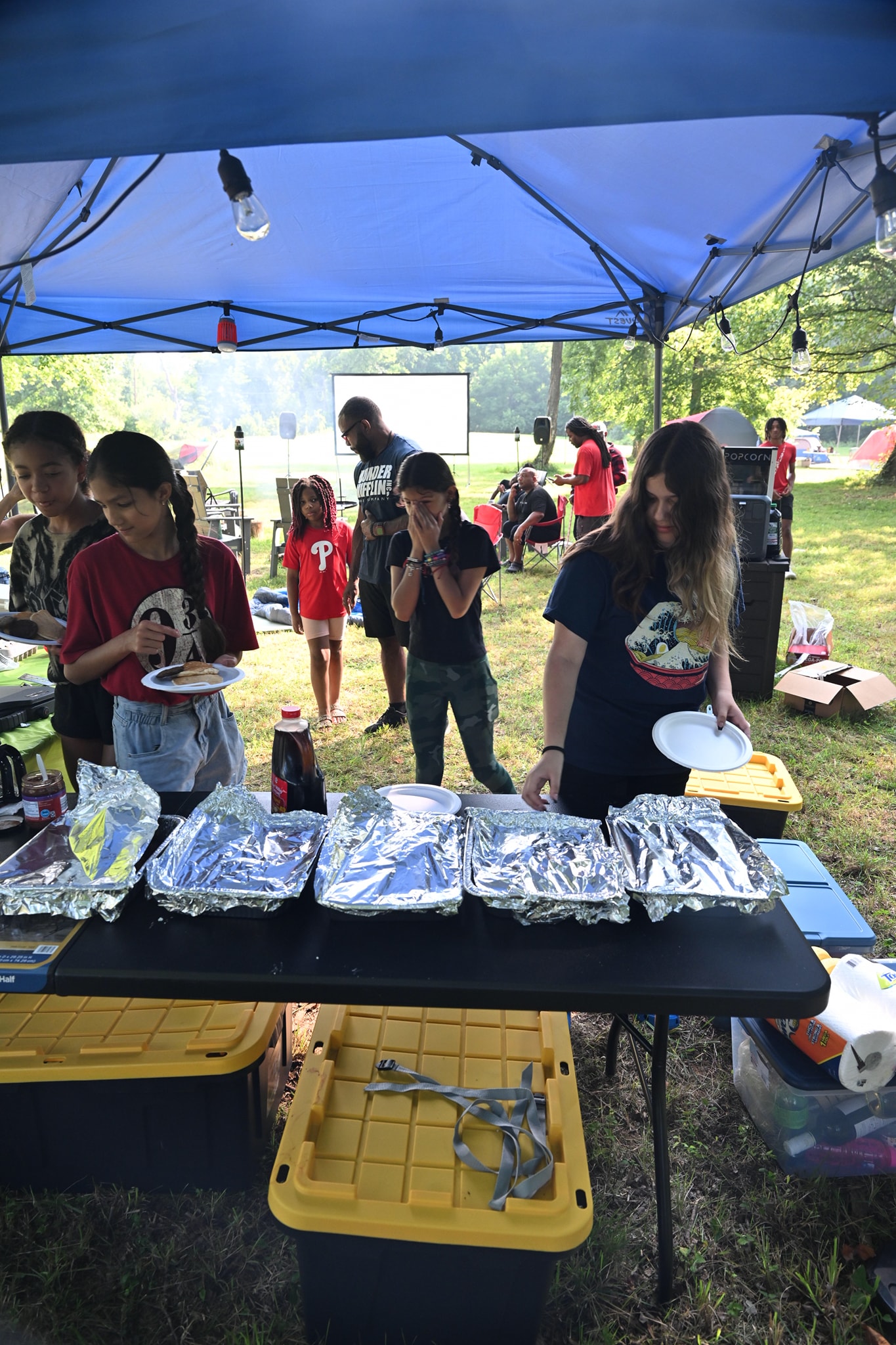 Kids filing through the food line under the blue canopy, loading up foil-covered trays — Campout 2023