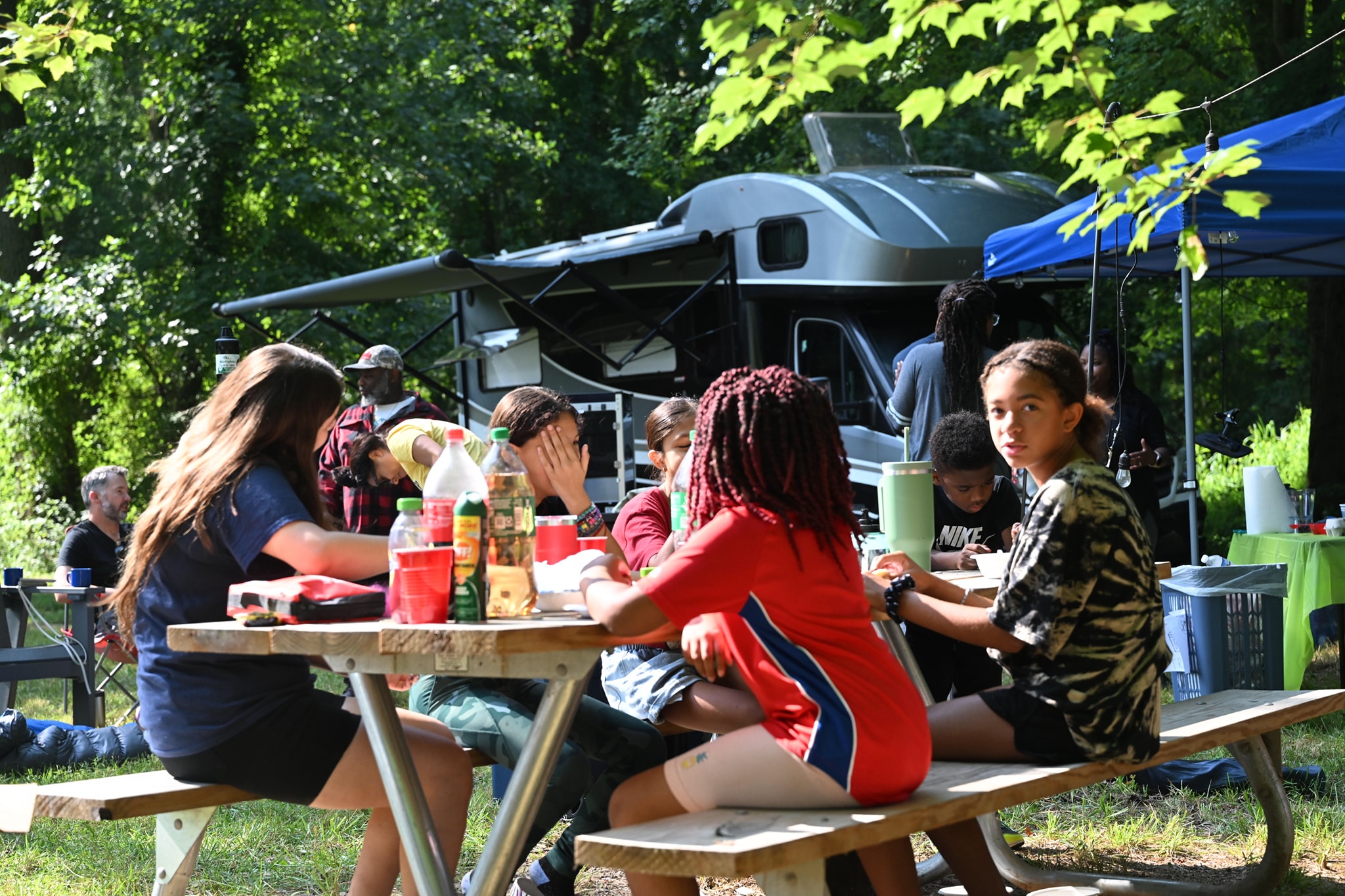 Kids eating at picnic tables in the shade with the RV and canopy visible behind — Campout 2023