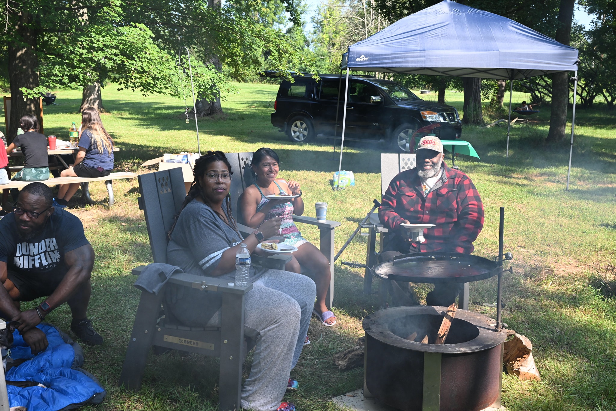 Guests eating beside the fire pit grill with a canopy tent and SUV in the background — Campout 2023