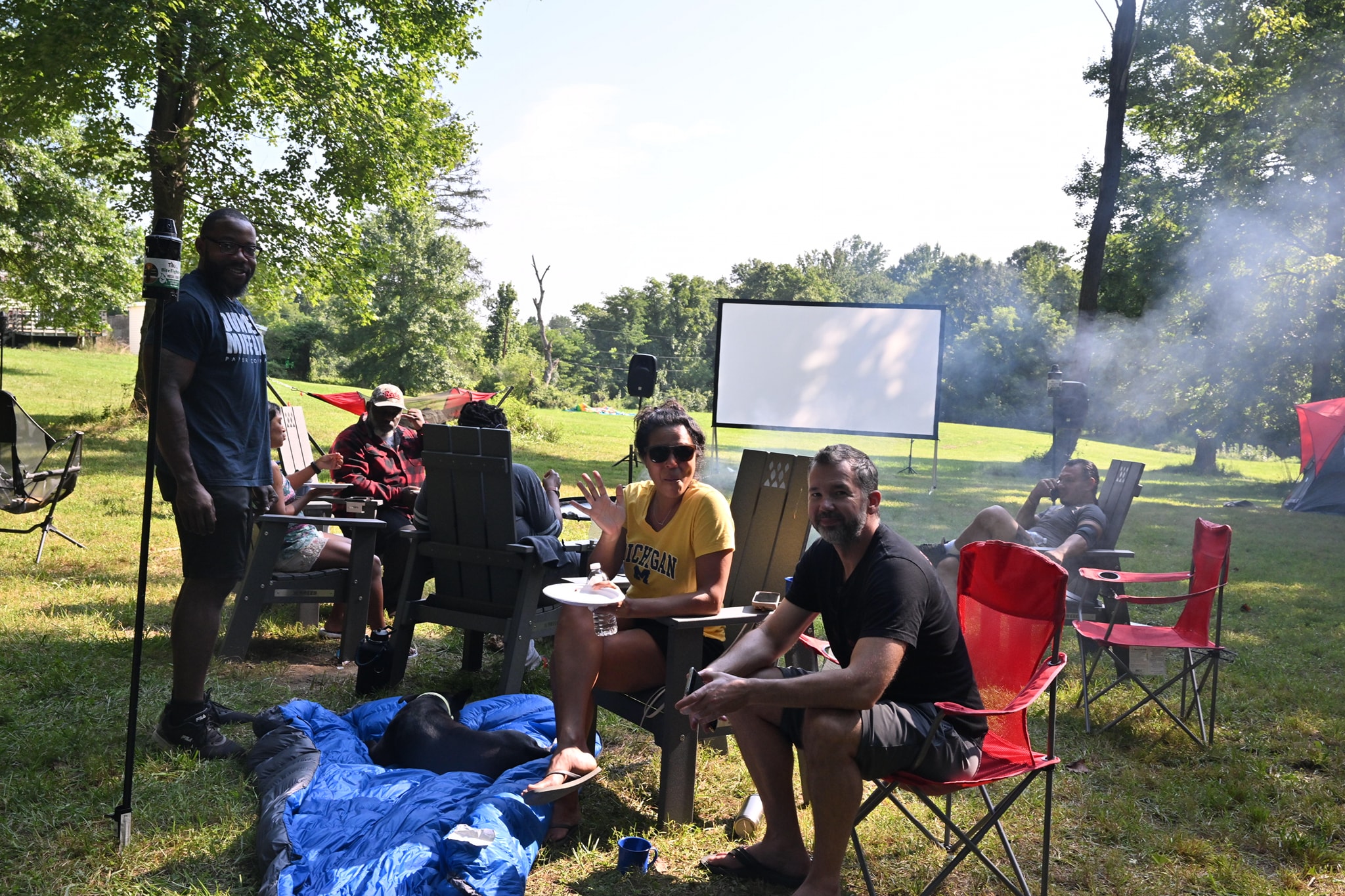 Guests relaxing in camp chairs in front of the movie screen with grill smoke drifting by — Campout 2023