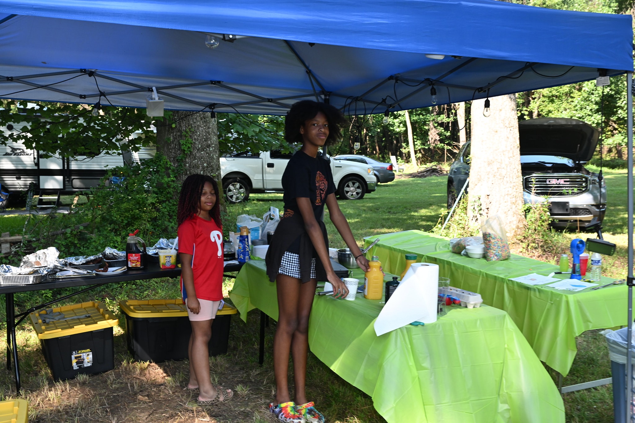 Two girls standing behind the food and drink tables under the blue canopy — Campout 2023