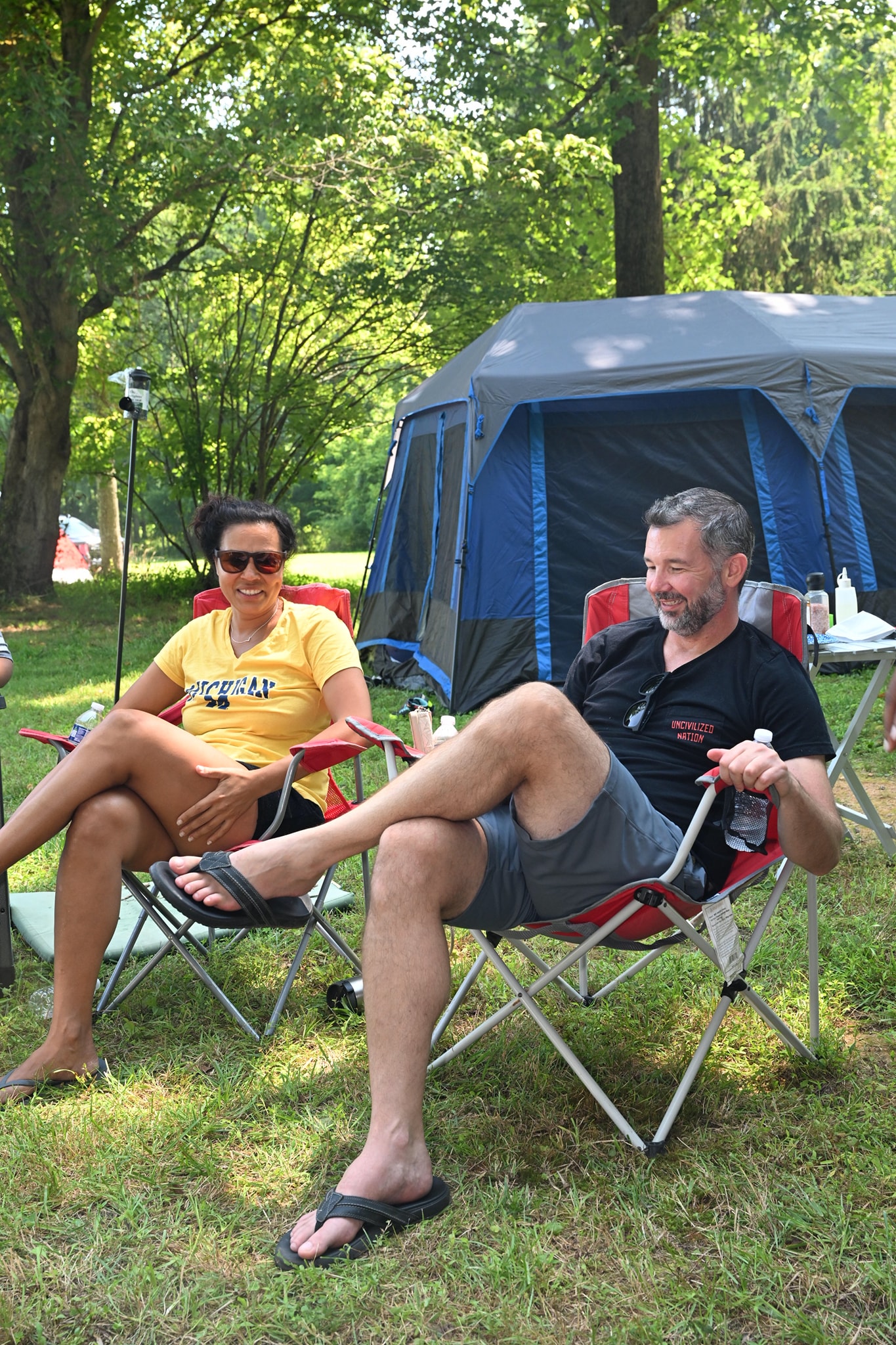 A couple laughing and relaxing in camp chairs in front of the large tent — Campout 2023
