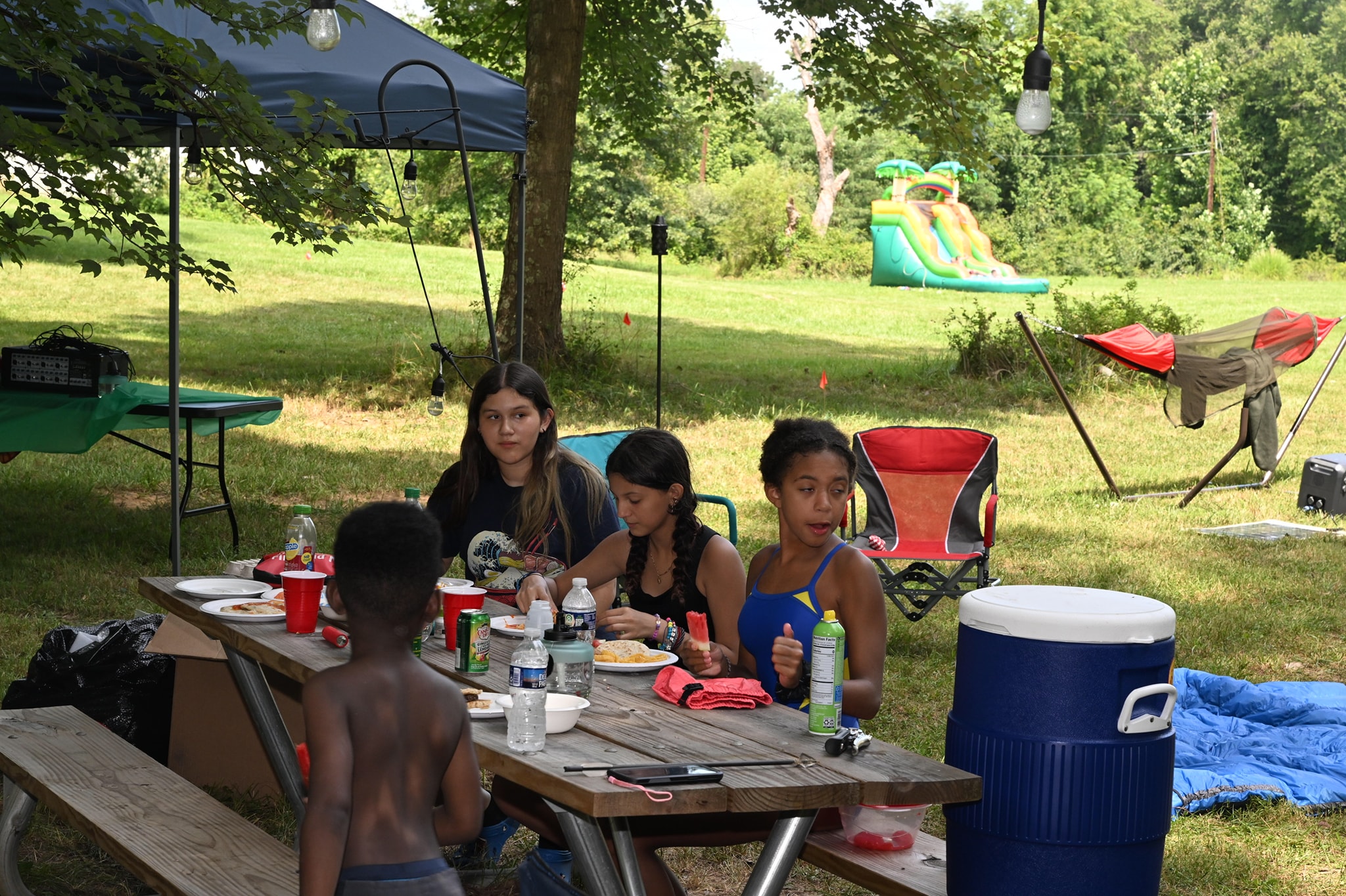 Kids eating at a picnic table under the canopy with the bounce house visible in the background — Campout 2023