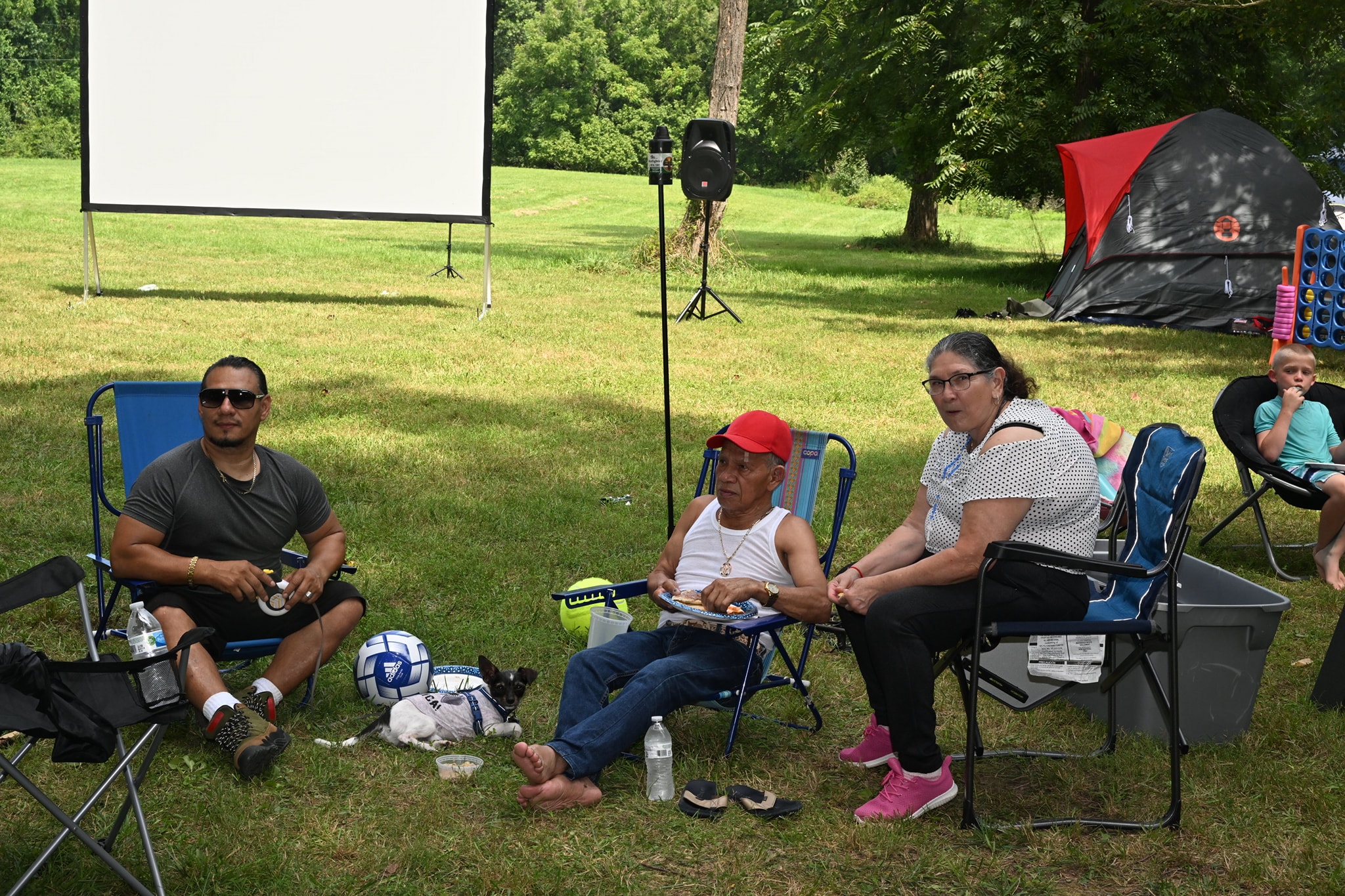Three guests relaxing in camp chairs in front of the movie screen with a soccer ball at their feet — Campout 2023