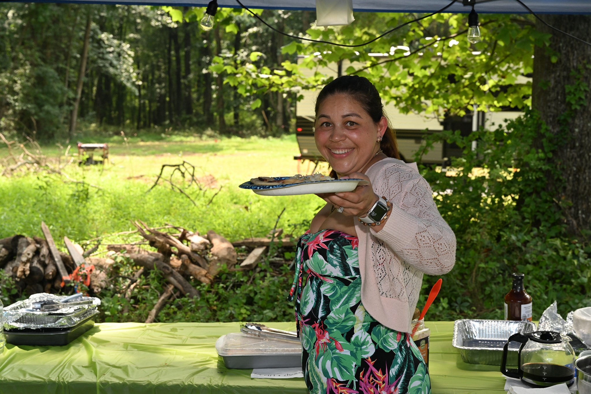 Smiling and holding out a plate at the food table under the canopy — Campout 2023