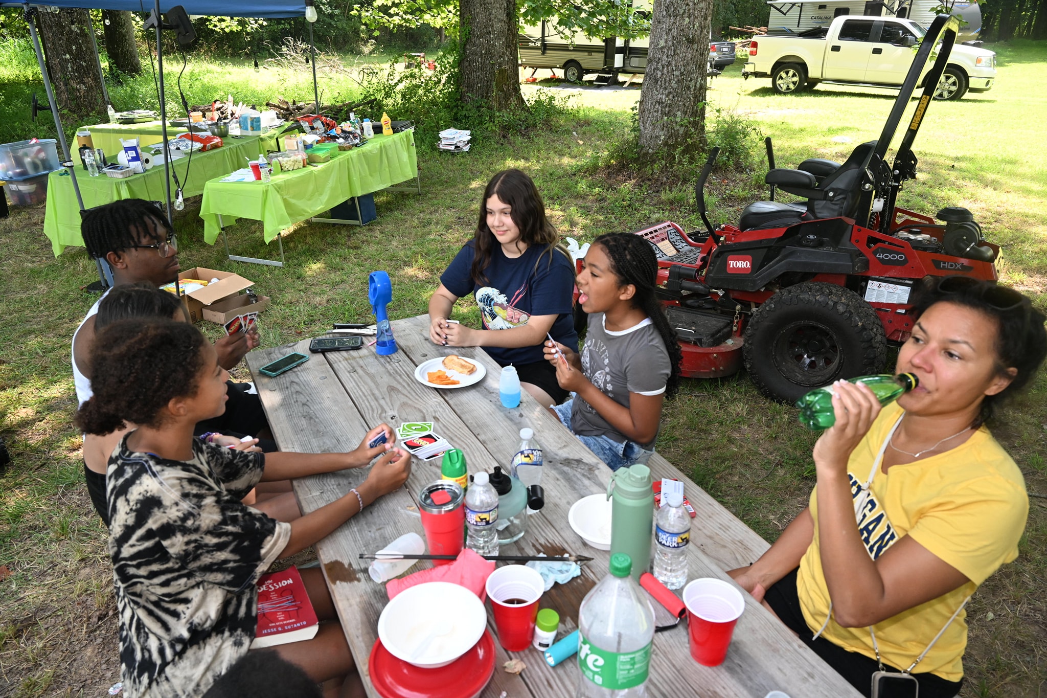 Guests and kids hanging out at a picnic table with food and drinks, the riding mower parked nearby — Campout 2023