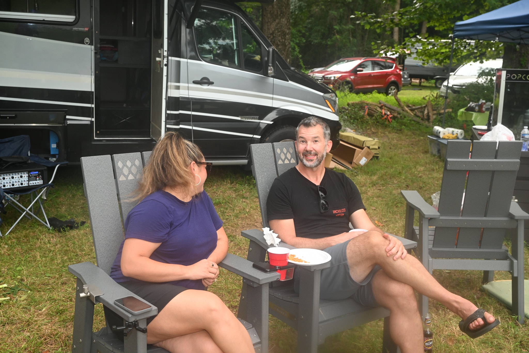 A couple chatting in Adirondack chairs beside the RV with food and drinks — Campout 2023
