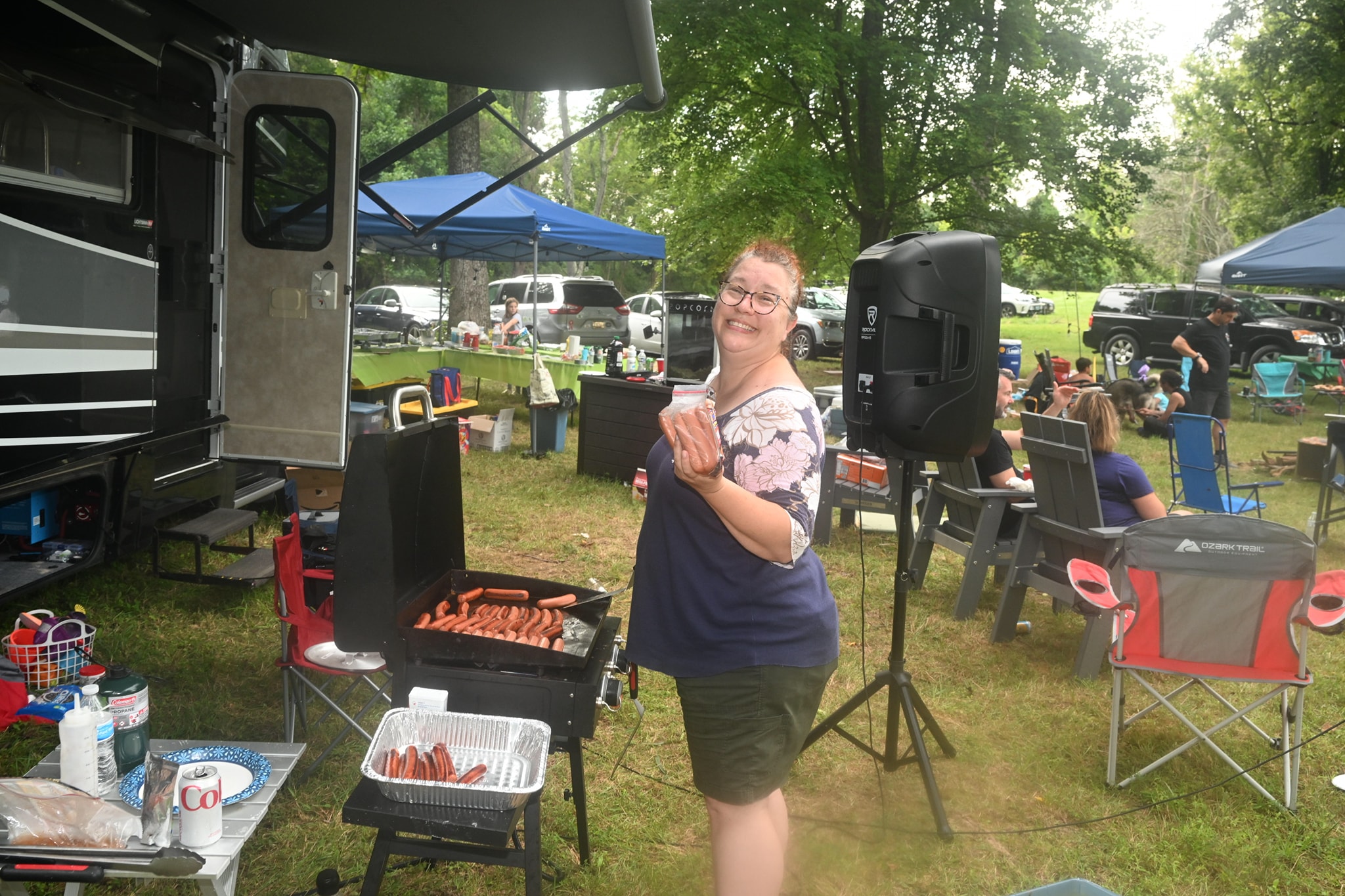Grilling hot dogs and smiling beside the speaker and RV with the full campsite behind — Campout 2023