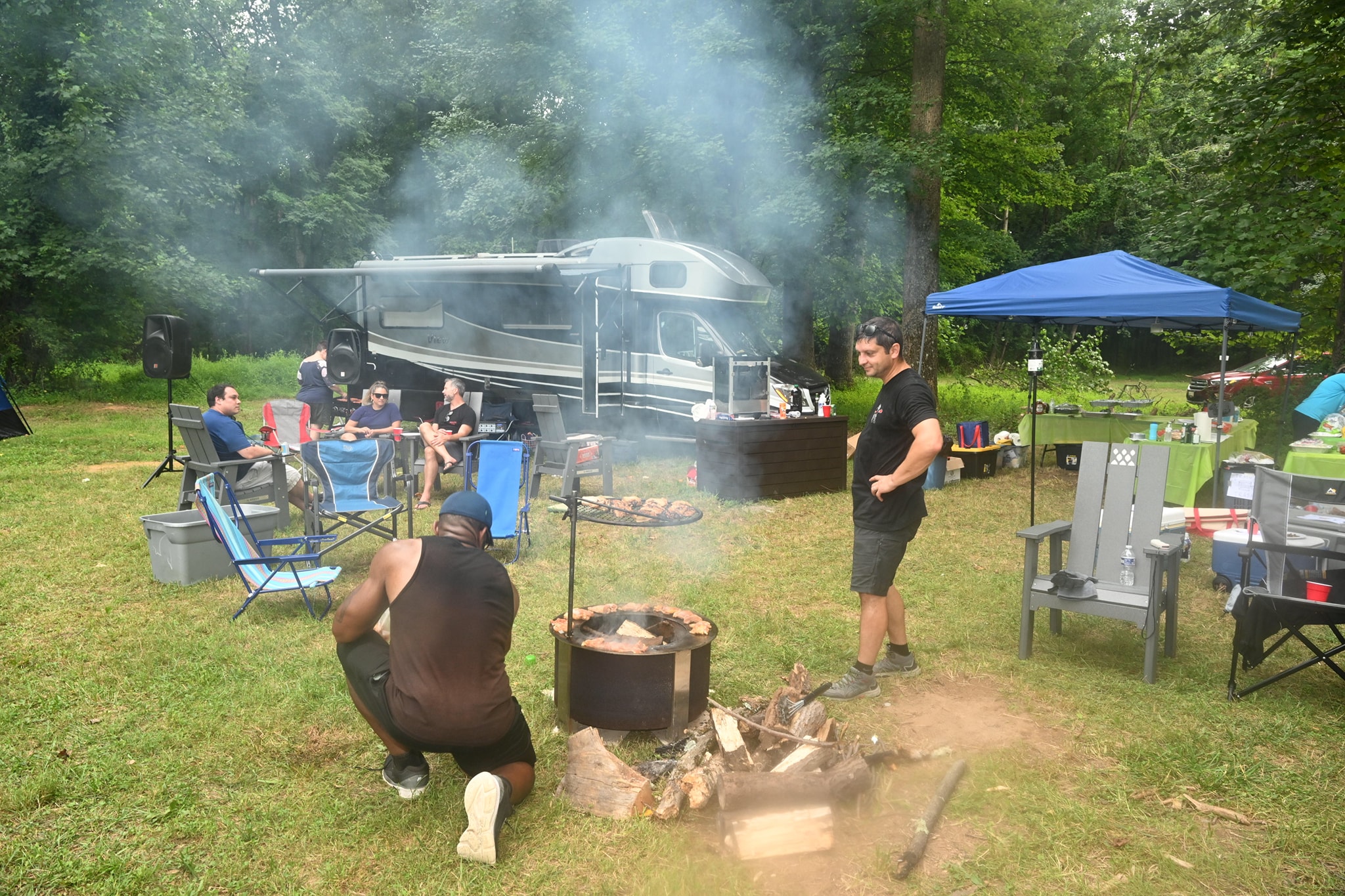 Two men tending the fire pit grill with smoke rising, the RV and blue canopy behind — Campout 2023