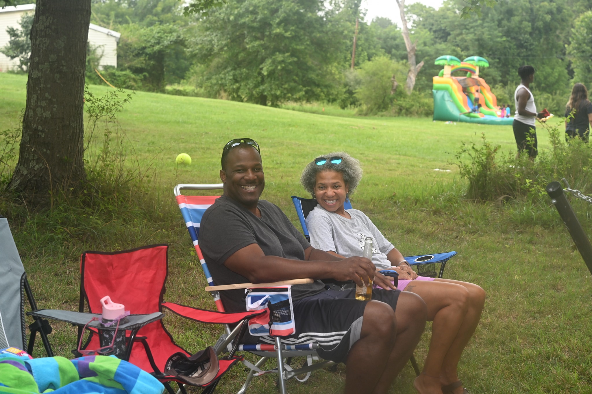 A couple relaxing in camp chairs with the bounce house and kids playing in the background — Campout 2023