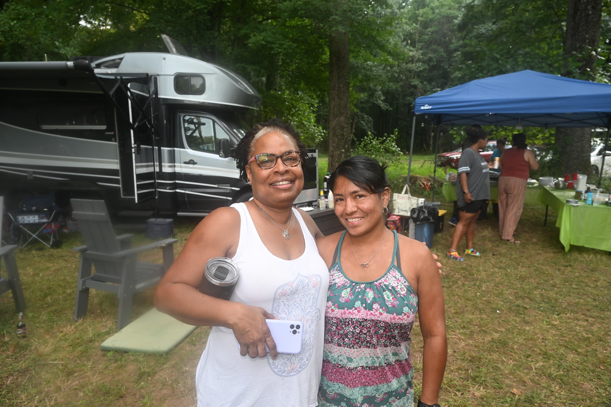 Two women smiling together in front of the RV and food tables — Campout 2023