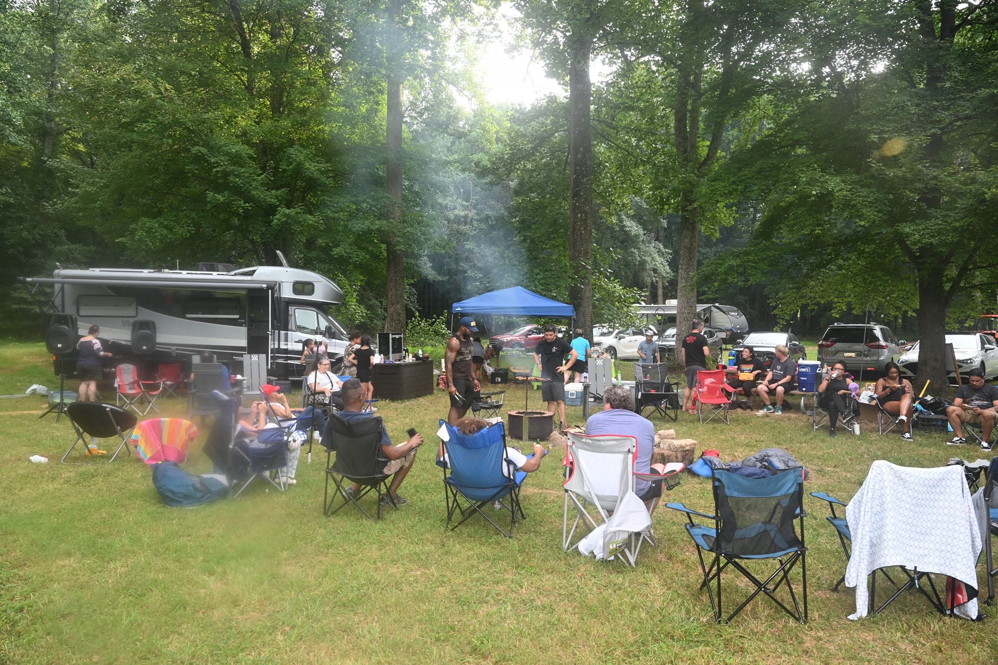 Wide view of the campsite with guests spread across camp chairs in the dappled shade, smoke rising from the grill — Campout 2023