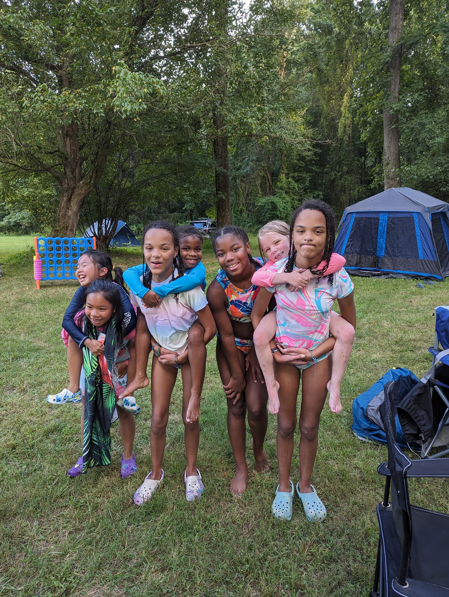A group of girls giving each other piggyback rides on the lawn with tents behind — Campout 2023
