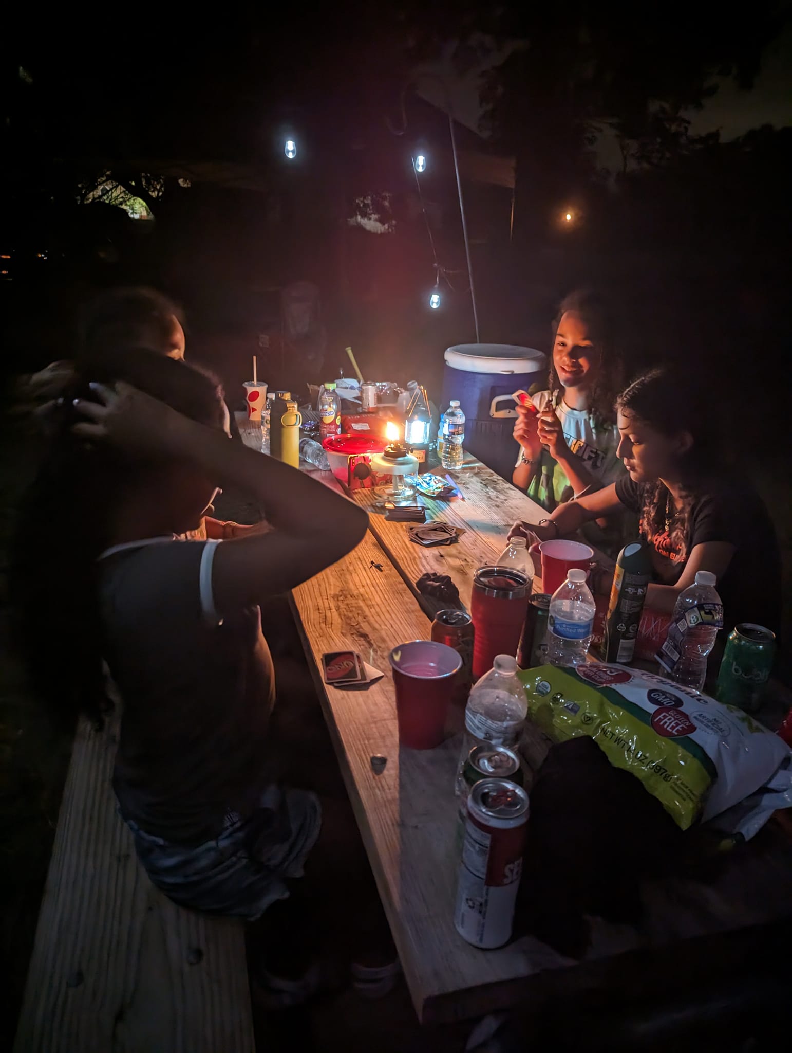 Guests laughing at a picnic table late at night under string lights with a lantern glowing — Campout 2023