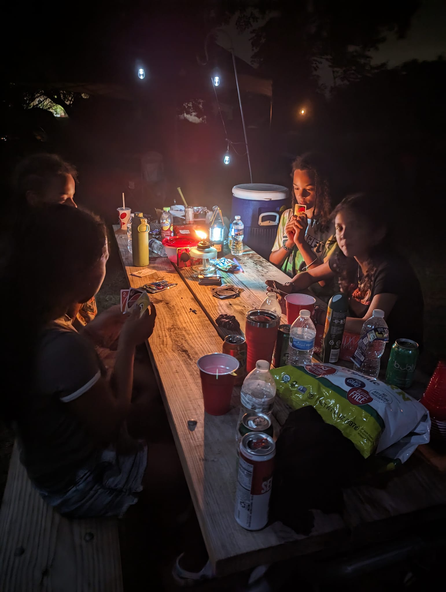 Kids and guests hanging out at the picnic table at night with snacks and drinks lit by a lantern — Campout 2023