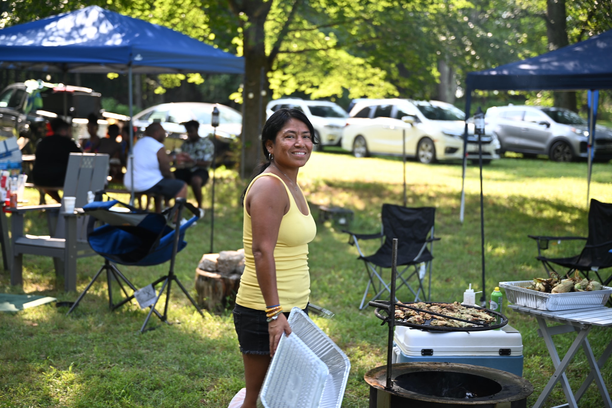 Smiling beside the grill in a yellow top with canopy tents behind — Campout 2024