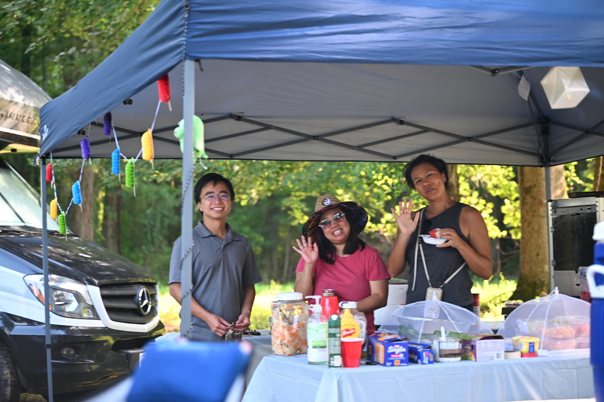 Guests waving from behind the food table under a colorful-flagged canopy — Campout 2024