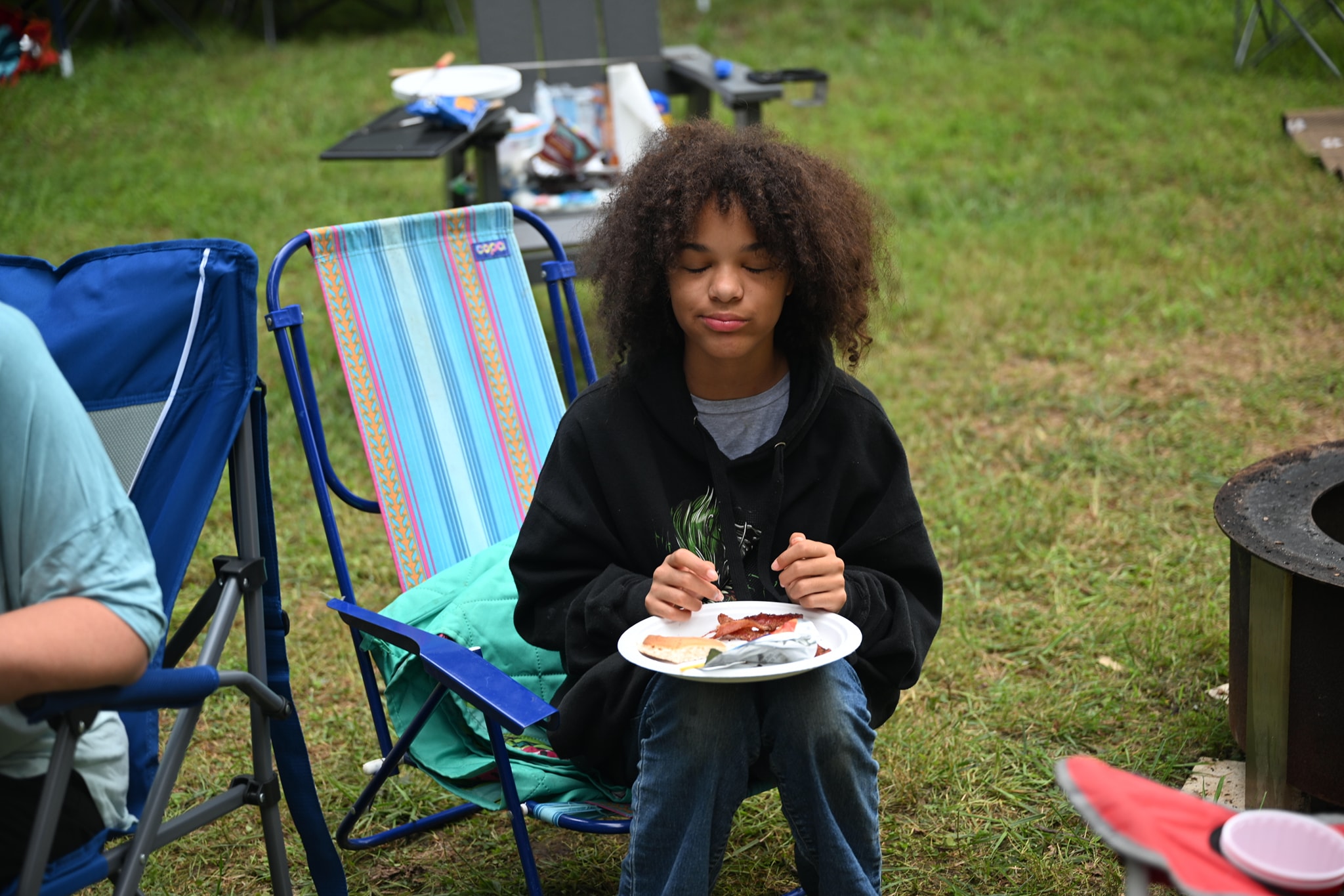 A kid eating from a plate in a camp chair beside the fire pit — Campout 2024