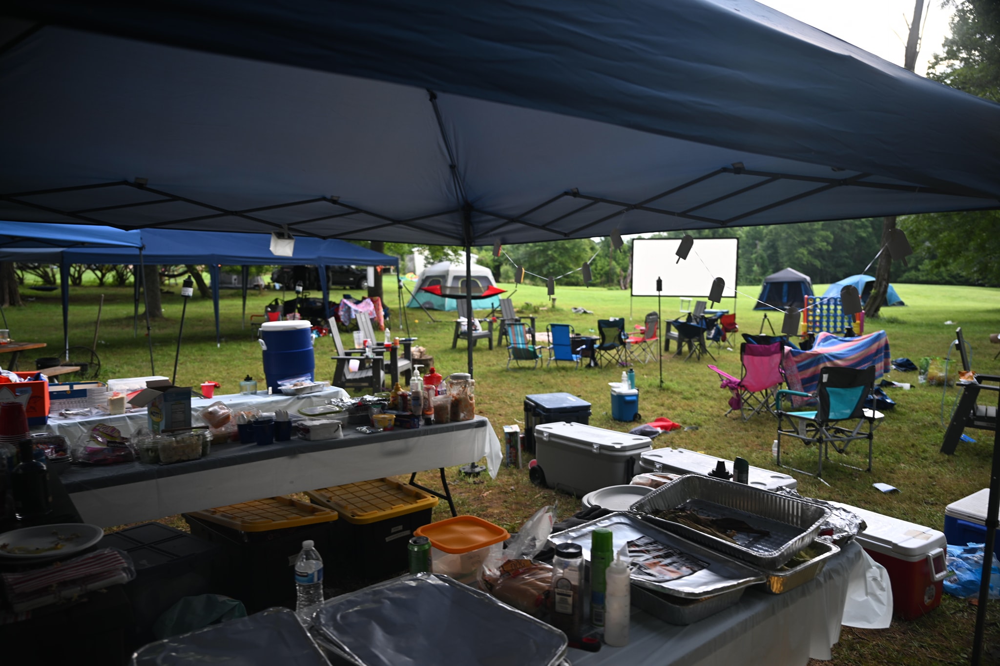 Food setup under the canopy with the movie screen and tents visible across the lawn — Campout 2024