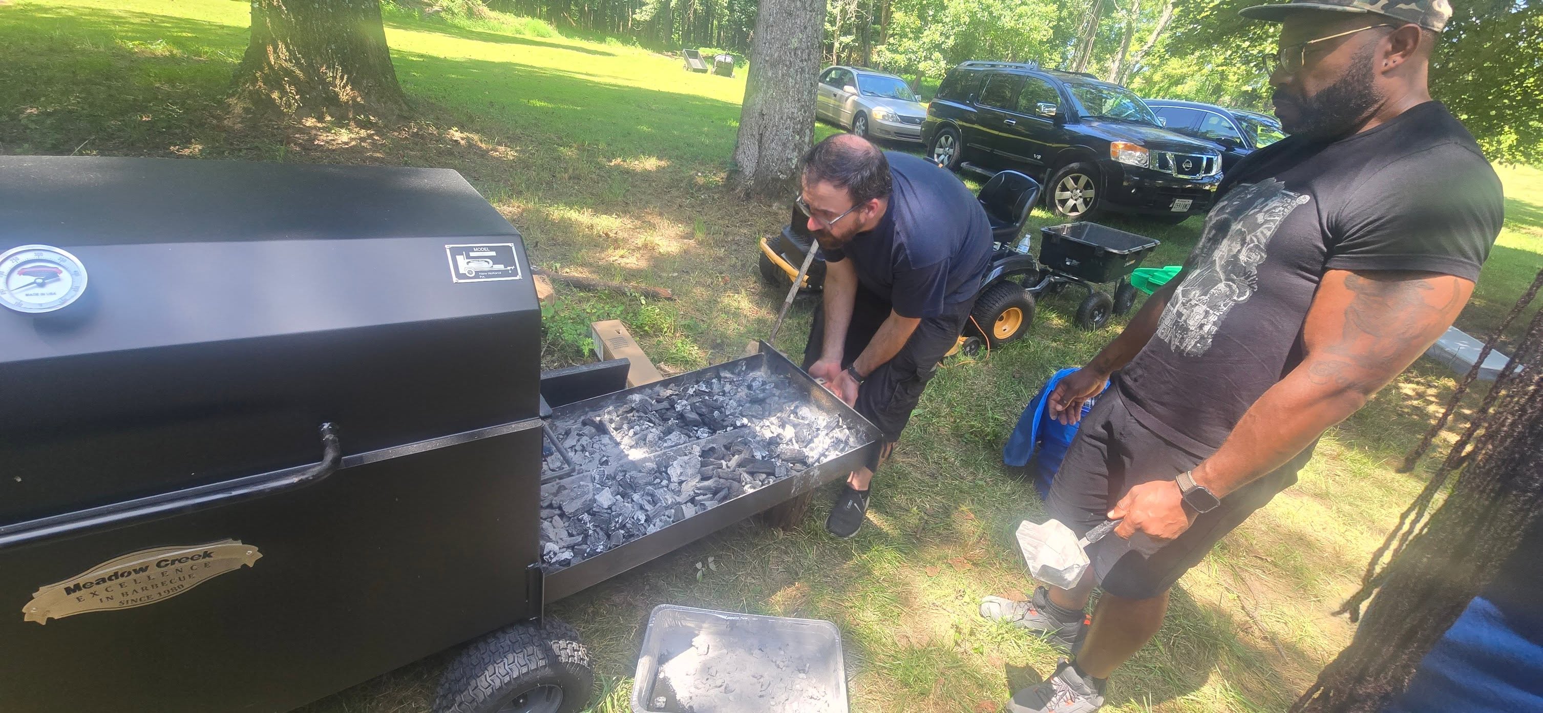 Loading charcoal into the smoker with the wooded yard behind — Campout 2025