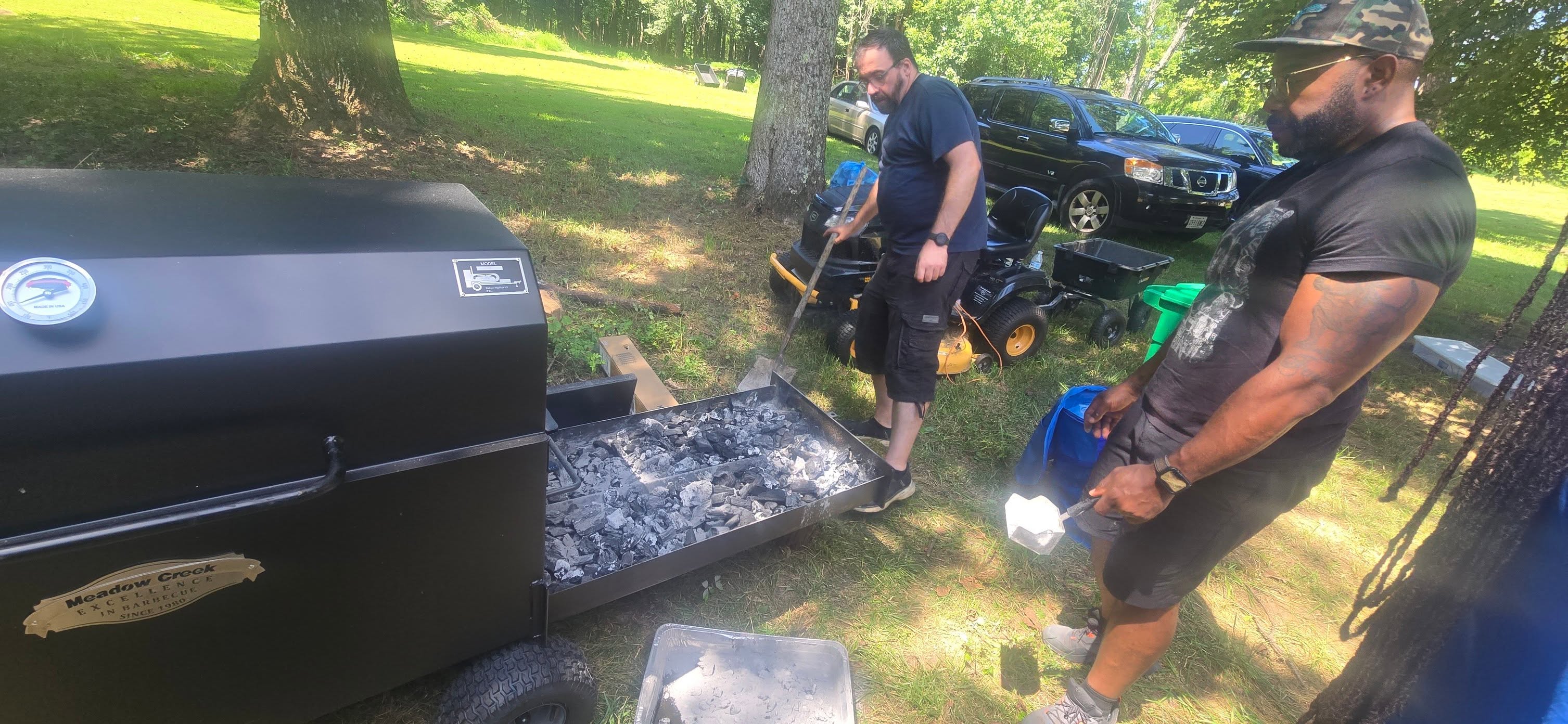 Managing the charcoal ash drawer on the smoker in the shade — Campout 2025