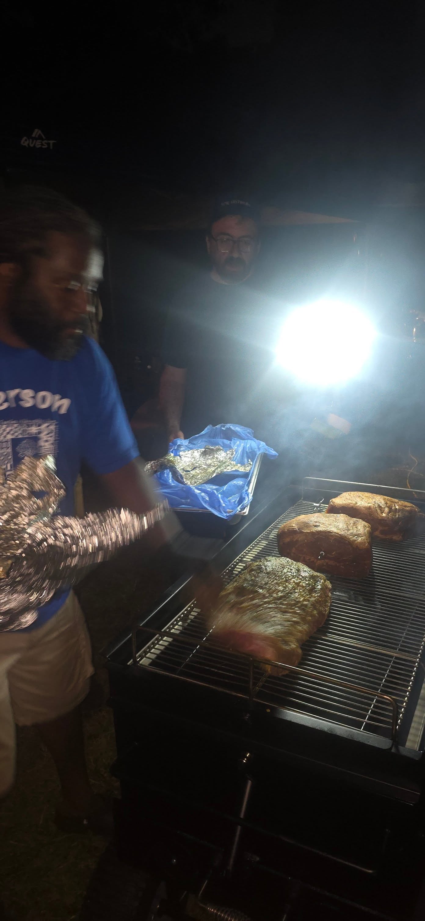 Pulling foil-wrapped brisket from the overnight smoker under work lights — Campout 2025