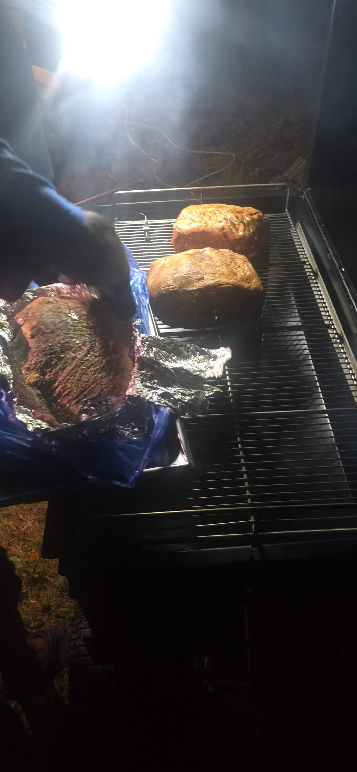 Gloved hand lifting brisket from the smoker rack late at night — Campout 2025
