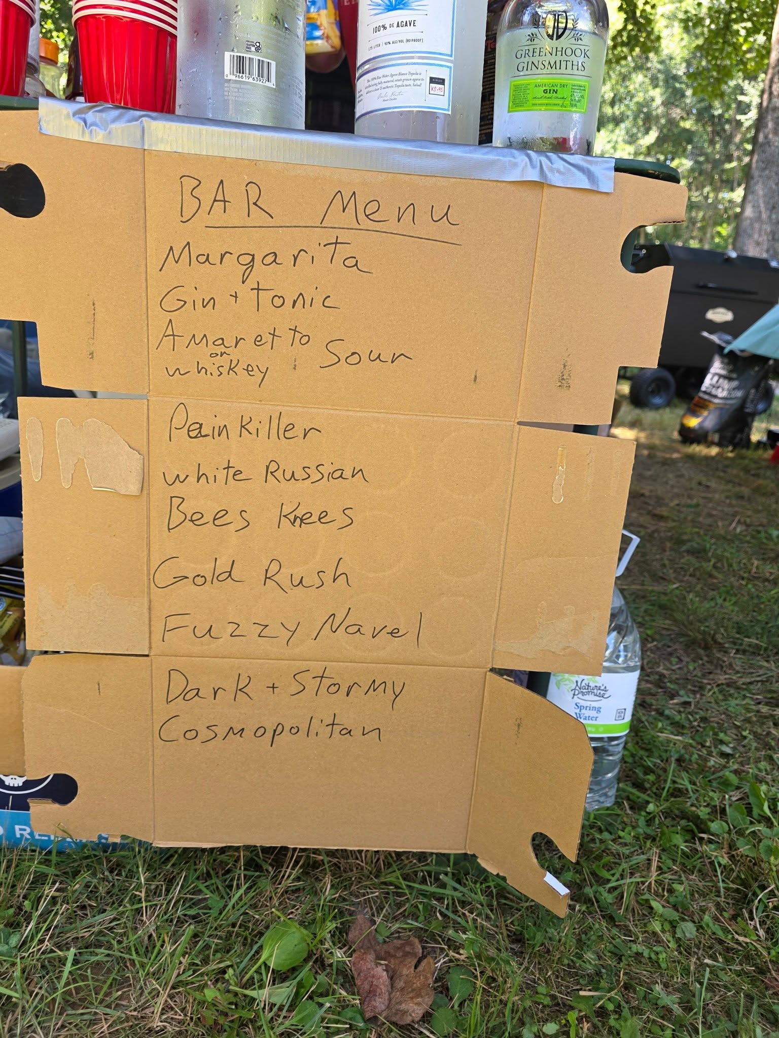 Handwritten bar menu on cardboard with ten cocktails and liquor bottles on top — Campout 2025