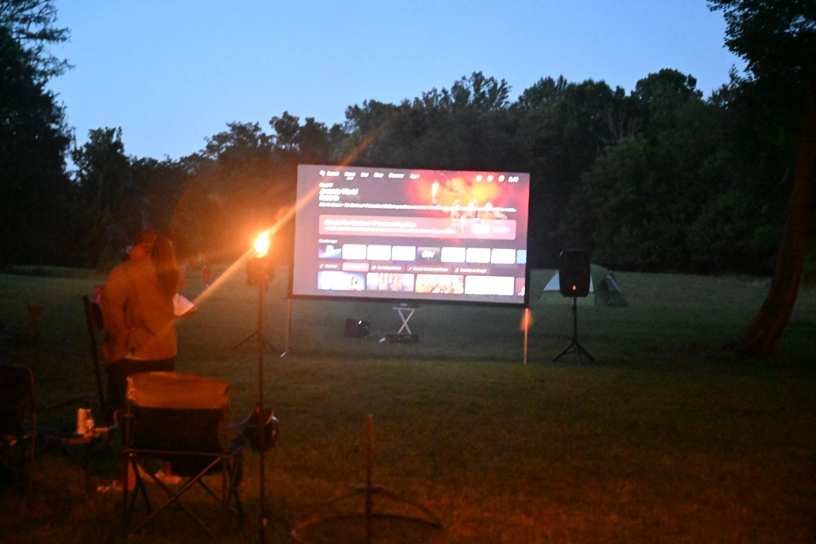 Setting up the outdoor movie screen at dusk, torches lighting the lawn — Campout 2025