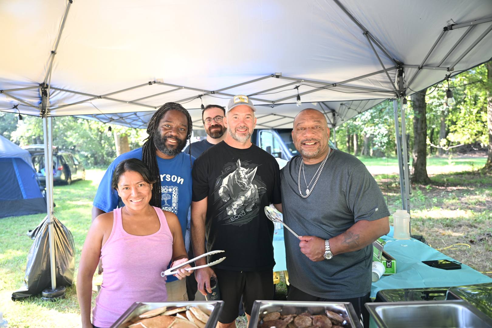 Smiling under the food canopy with serving tongs and trays in hand — Campout 2025