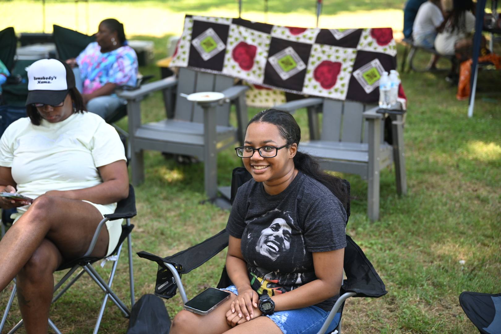 Smiling in a camp chair with a colorful quilt on the bench behind — Campout 2025
