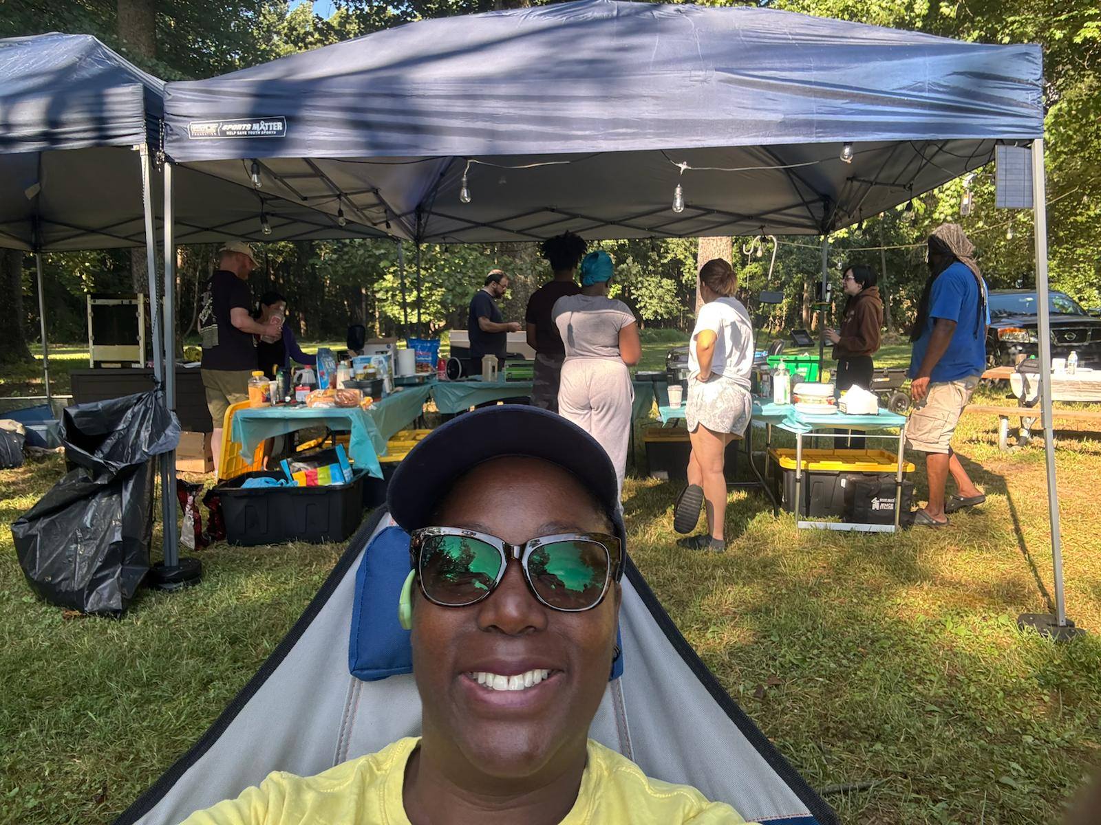 A selfie from a camp chair with the catering crew setting up behind — Campout 2025